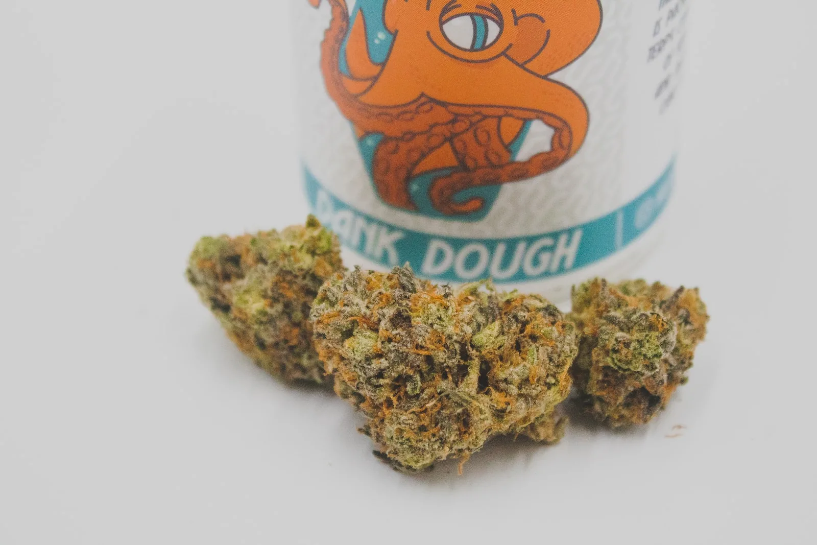 Close-up of cannabis buds in front of a container labeled "Dank Dough," featuring an octopus design. The image highlights the vibrant green and orange hues of the buds, showcasing their texture and quality.