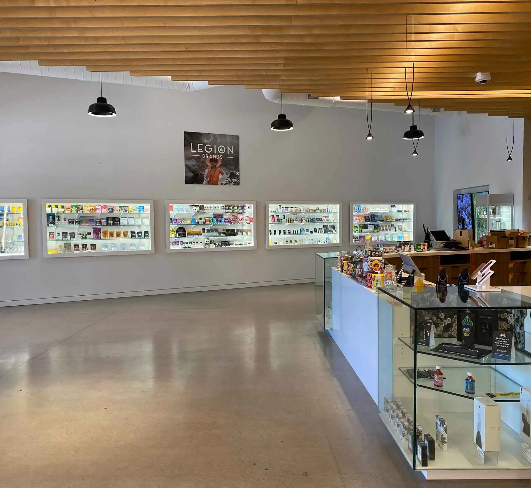 Interior view of a modern retail store showcasing multiple display cases filled with various products, including vaping supplies and accessories. The store features a sleek design with wooden ceiling panels and stylish lighting, promoting a welcoming atmosphere. A prominent sign for "Legion Brands" is displayed on the wall, enhancing brand visibility.